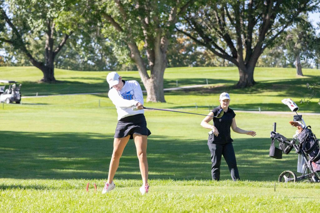 A female golfer swings a club on a sunny day while another woman watches nearby, with trees and a golf cart in the background.