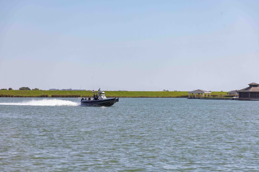A black boat speeding across a calm body of water, with green grass and structures in the background.