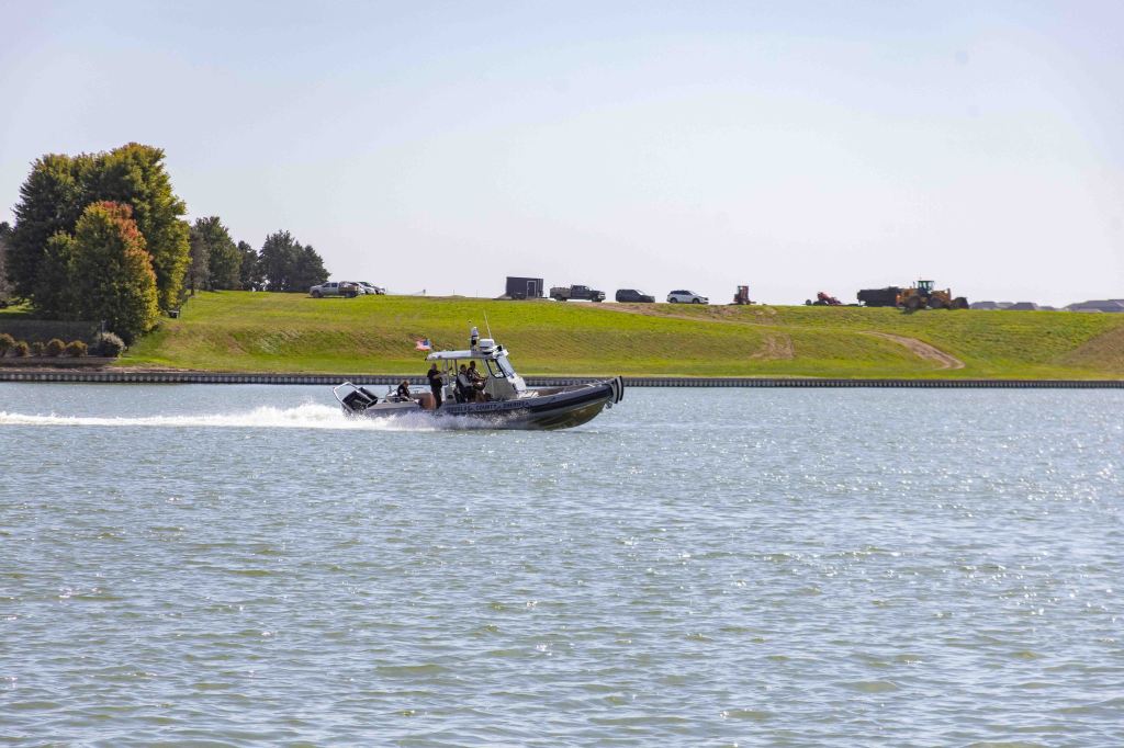 A patrol boat moving swiftly on the water with a green hillside and vehicles in the background.