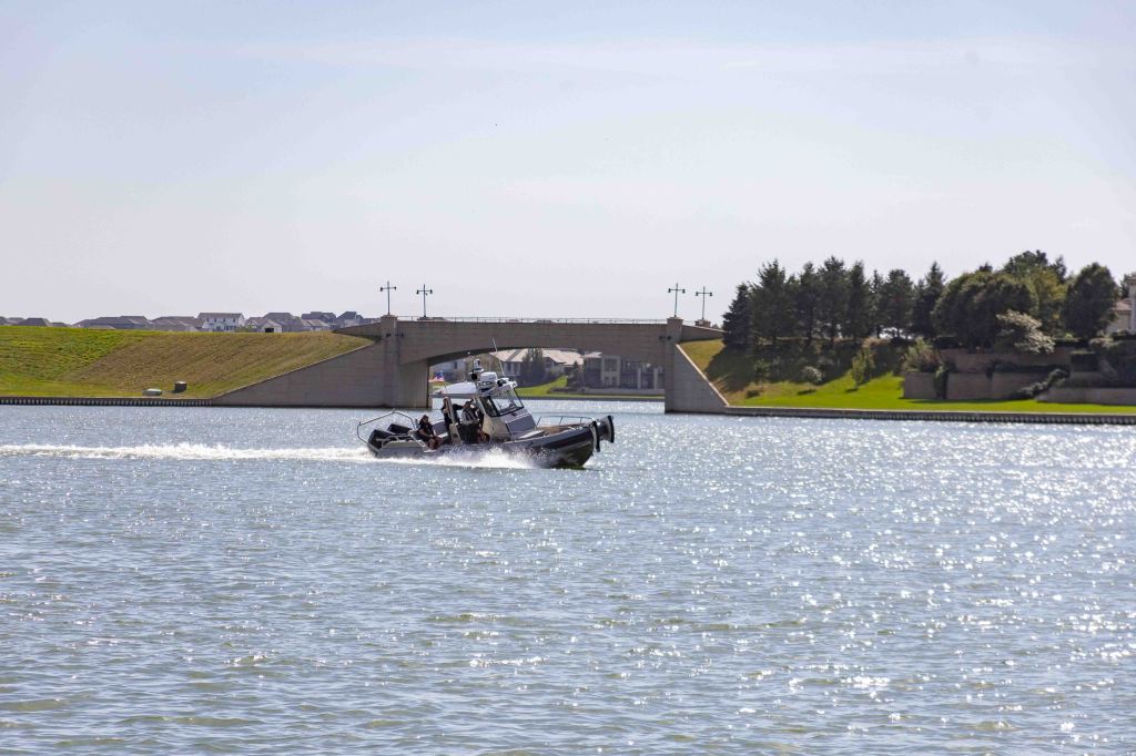 A motorboat navigates through a body of water near a bridge, with green hills and trees in the background.
