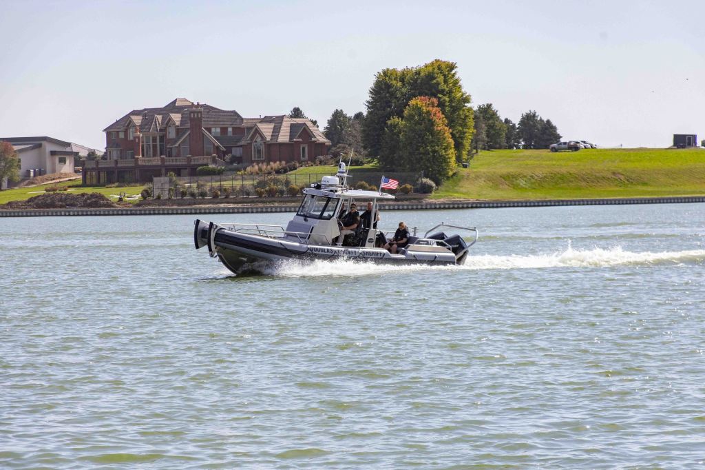 A police boat is cruising on a lake with a large house and green landscape in the background.