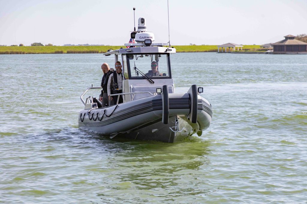 A small gray boat navigates through calm green waters, with two men on board wearing life jackets and sunglasses.