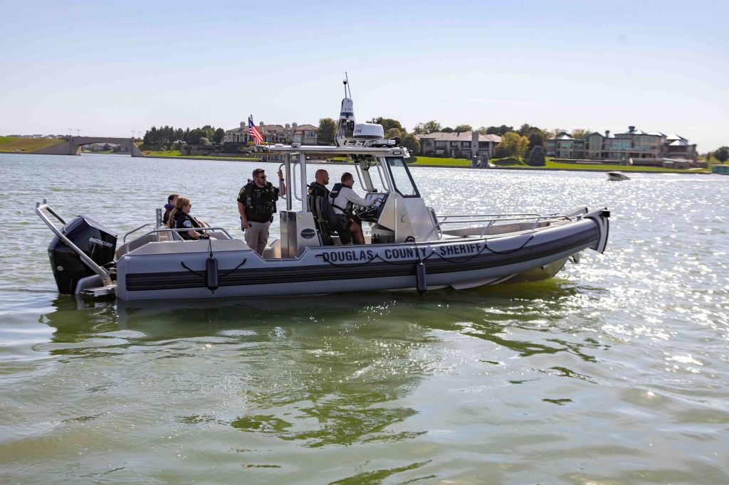A Douglas County Sheriff boat with law enforcement officers on board patrols a calm water body on a sunny day.