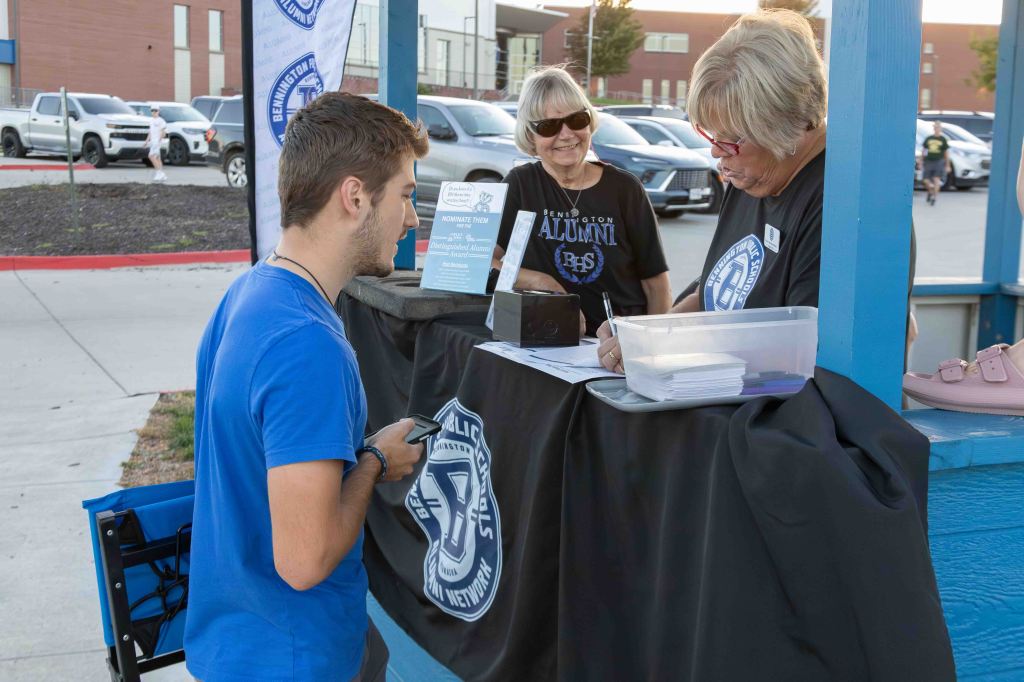 A young man in a blue shirt is speaking with two women at a registration table for an alumni event, while a few cars are parked in the background.