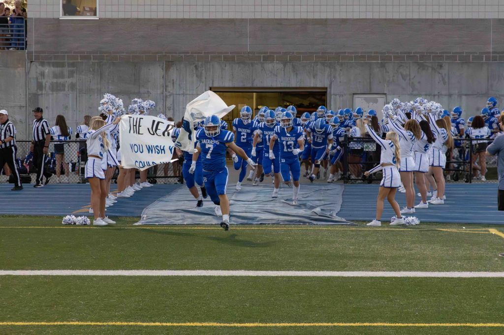 High school football team running onto the field through a paper banner, flanked by cheerleaders. The team wears blue uniforms and helmets, while cheerleaders wear white and blue outfits.