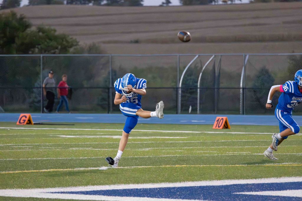 A high school football player in a blue uniform kicks a football on the field during a game, with teammates and spectators in the background.