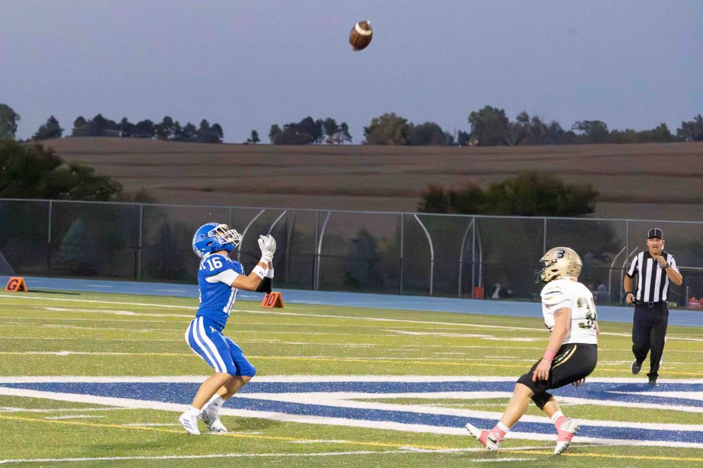 A football player in a blue uniform, number 16, catches a pass during a game, with an opposing player in a white uniform nearby and an official observing the play.