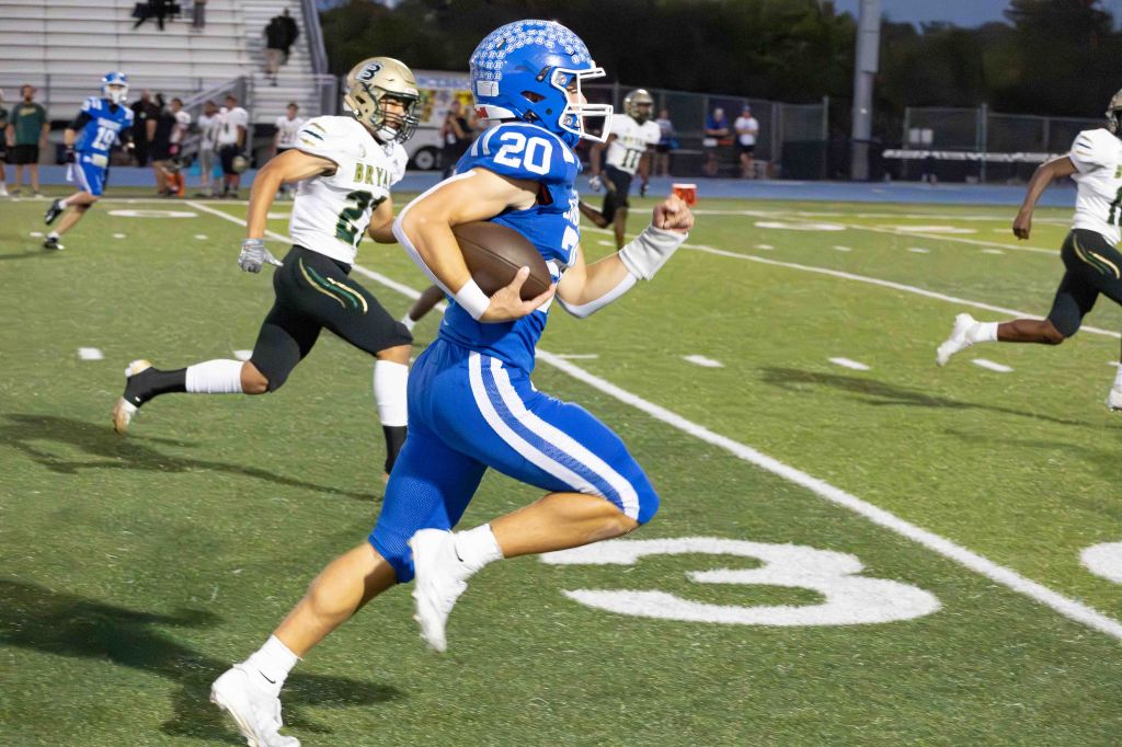 A football player in a blue jersey with the number 20 runs with the ball on the field, while players in black and gold chase from behind.