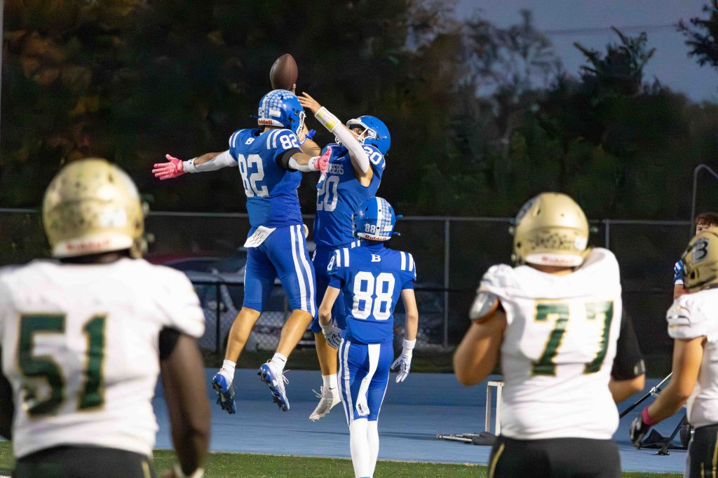 Two football players in blue jerseys celebrate after a successful catch, while players in white and gold watch from the foreground.