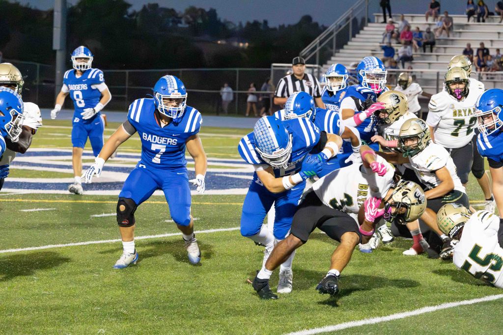 High school football game action between the Badgers and an opposing team, featuring players in blue and white uniforms tackling a player in black and gold.