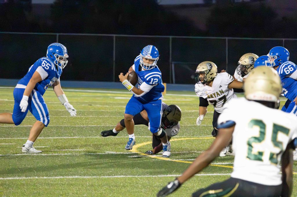 A high school football game scene showing a player in blue jersey number 12 running with the ball while being pursued by defenders in black and gold uniforms on a well-lit field.