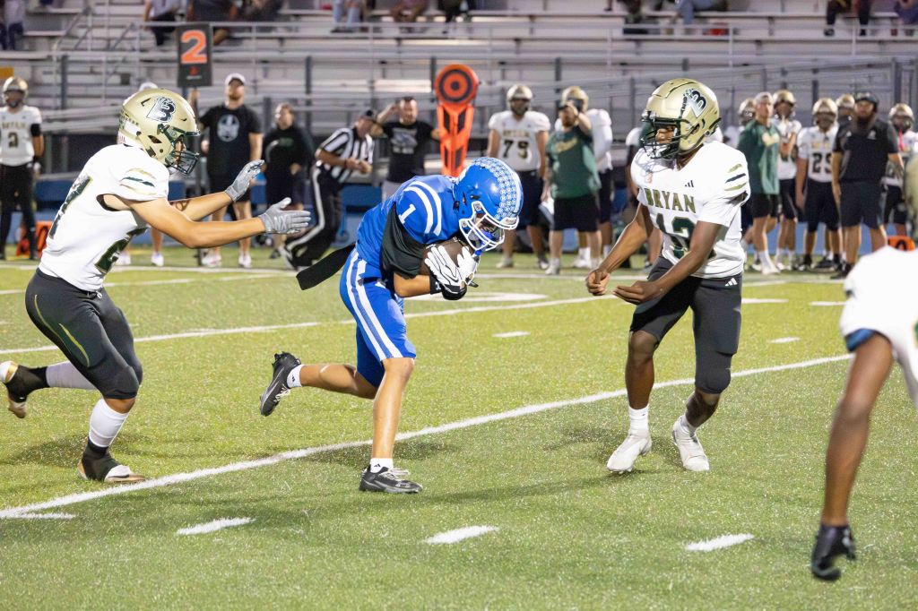 A youth football game in progress, featuring two teams. One player in a blue jersey is running with the ball, while two opponents in black and white jerseys attempt to tackle him.
