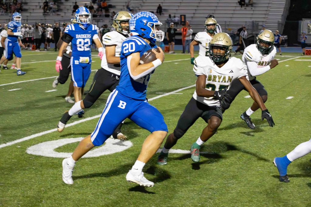 A football player in a blue jersey with the number 20 is running with the ball on the field, evading two defenders in white and gold uniforms.