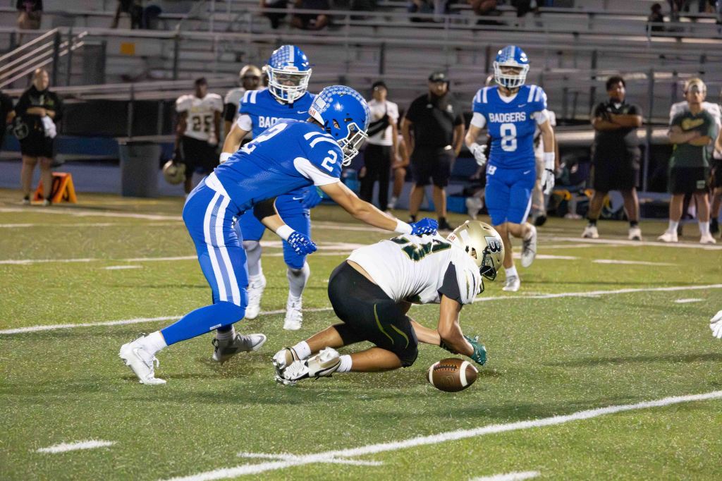 A high school football game featuring two teams, with a player in a blue uniform tackling an opponent in a white uniform who is attempting to recover a loose football on the field.