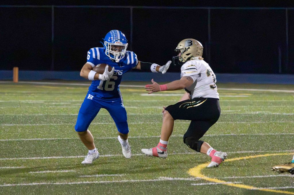 A football player in a blue uniform with the number 16 dodges an opponent in a gold and black uniform on a football field.