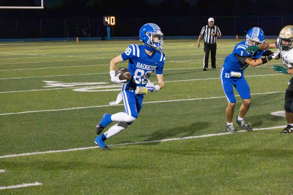 A football player wearing a blue uniform with the word 'BADGERS' running with the ball during a game, while another player in the background attempts to tackle.