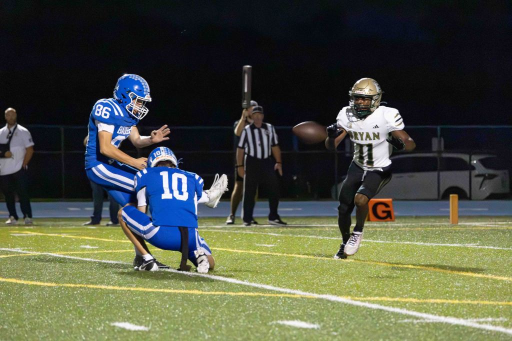 A high school football game scene featuring a player in a blue jersey preparing to kick while another player in the same team holds the ball. An opposing player in a white and gold jersey is evading a defender in the background as he runs with the football.