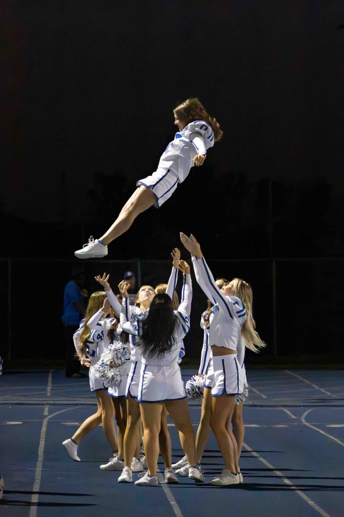 A cheerleader being tossed into the air by a group of teammates during a night performance, with a blue track visible in the background.