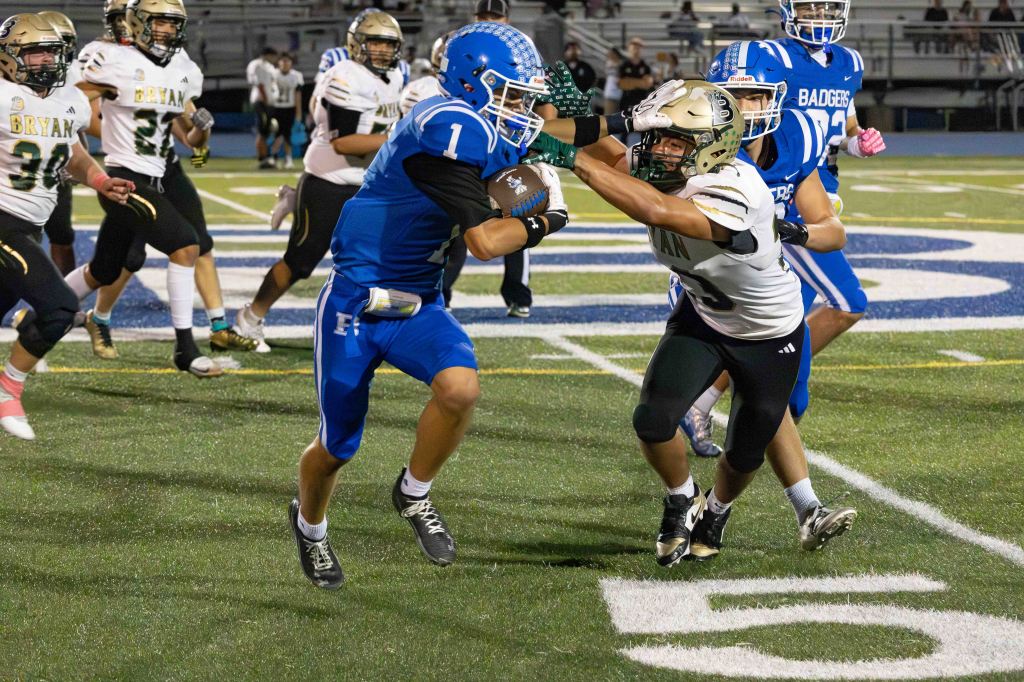 A high school football game scene, featuring a player in blue uniform attempting to evade a tackle from a player in white uniform, while other players from both teams are engaged in the background.