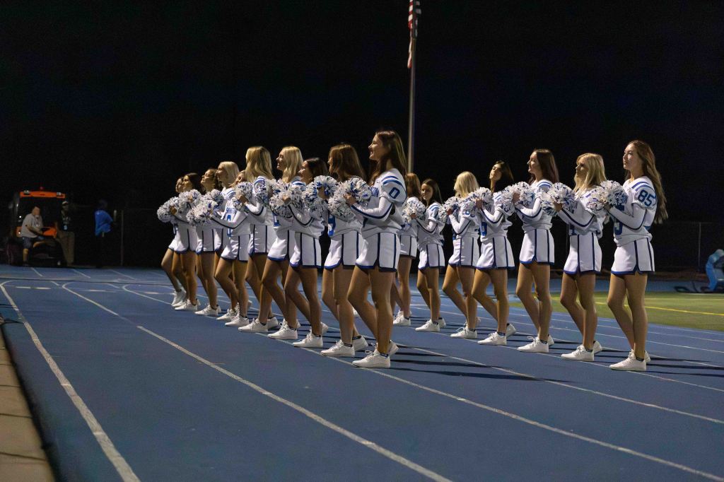 A group of cheerleaders in matching uniforms performs on a track during a night event, holding pom-poms and displaying synchronized movements.