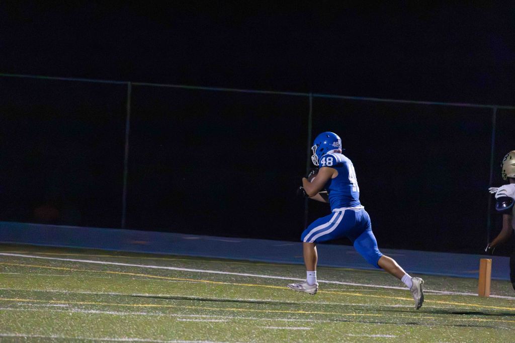 A football player in a blue uniform, number 48, running on a field at night with the football in hand.