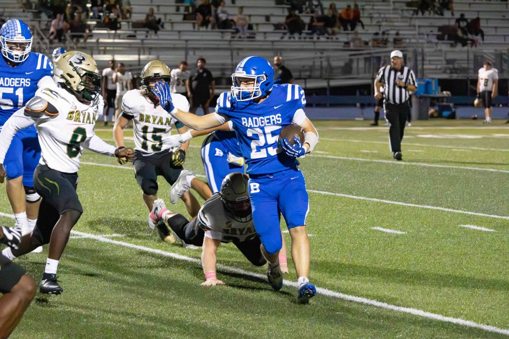 A football player in blue and white jersey number 25 runs down the field while being pursued by opponents in black and white jerseys during a night game.
