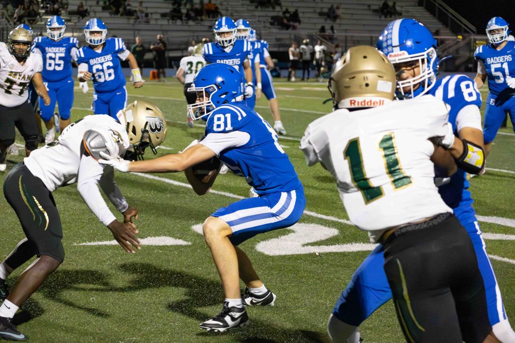A high school football game in progress, featuring players in blue and gold uniforms engaging in a tackle. One player wearing blue number 81 is attempting to break free while facing off against opponents in white and gold helmets.