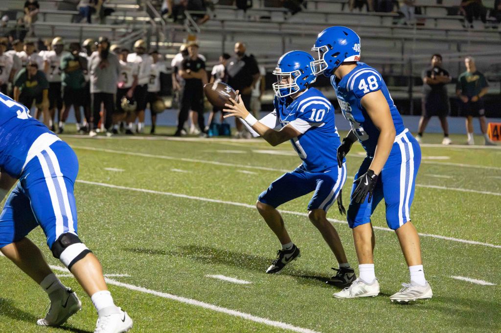 A young football player in a blue uniform prepares to throw the ball while another player stands ready nearby, during a game at night.