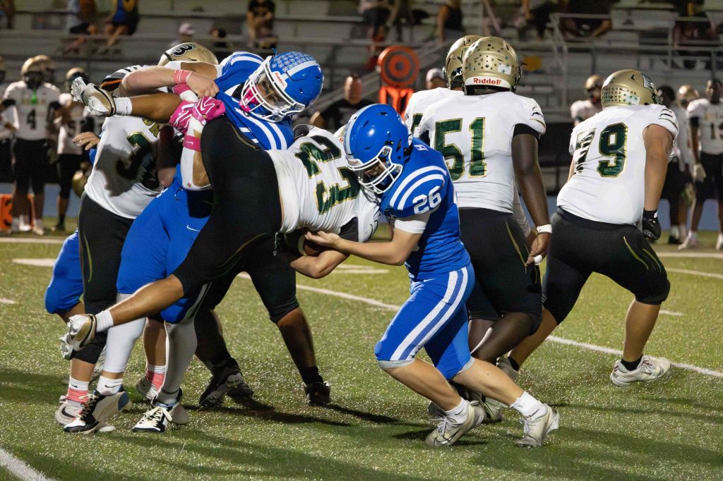 A high school football game in progress, featuring players in blue and gold uniforms engaged in a tackle on the field.