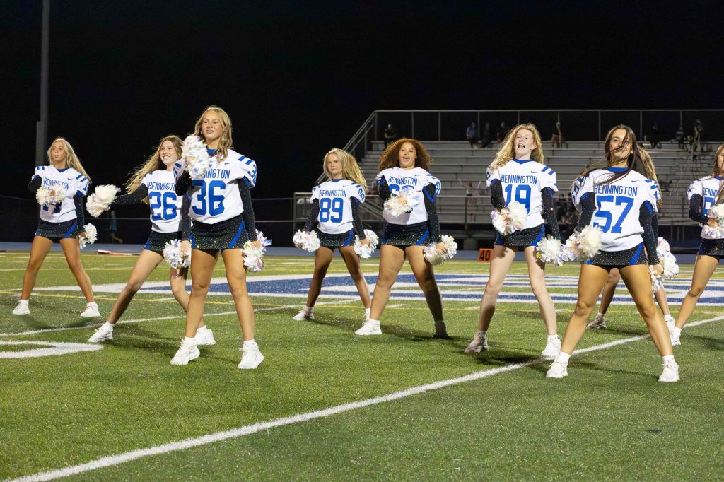 A group of cheerleaders performing at a football game under the night sky, wearing blue and white uniforms with pom poms.
