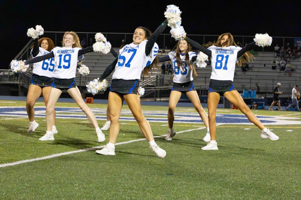 A group of five cheerleaders performing on a football field at night, wearing white jerseys with blue accents and pom-poms.