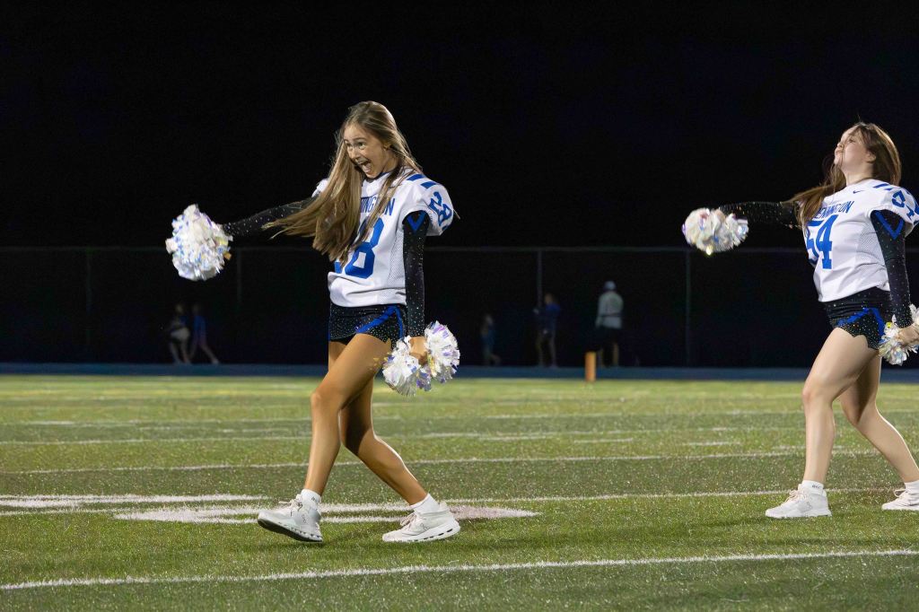 Two cheerleaders performing on a football field at night, one is energetically cheering with pom-poms and smiling, while the other is also in motion.