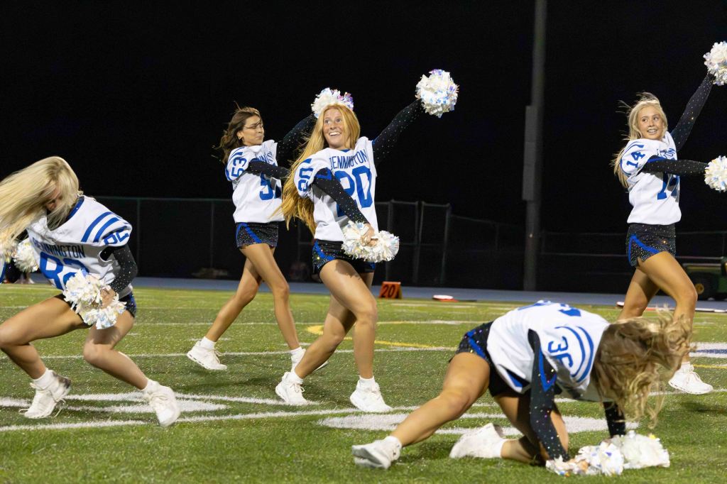 A group of cheerleaders performing energetically on a football field at night, wearing sporty outfits and holding pom-poms.
