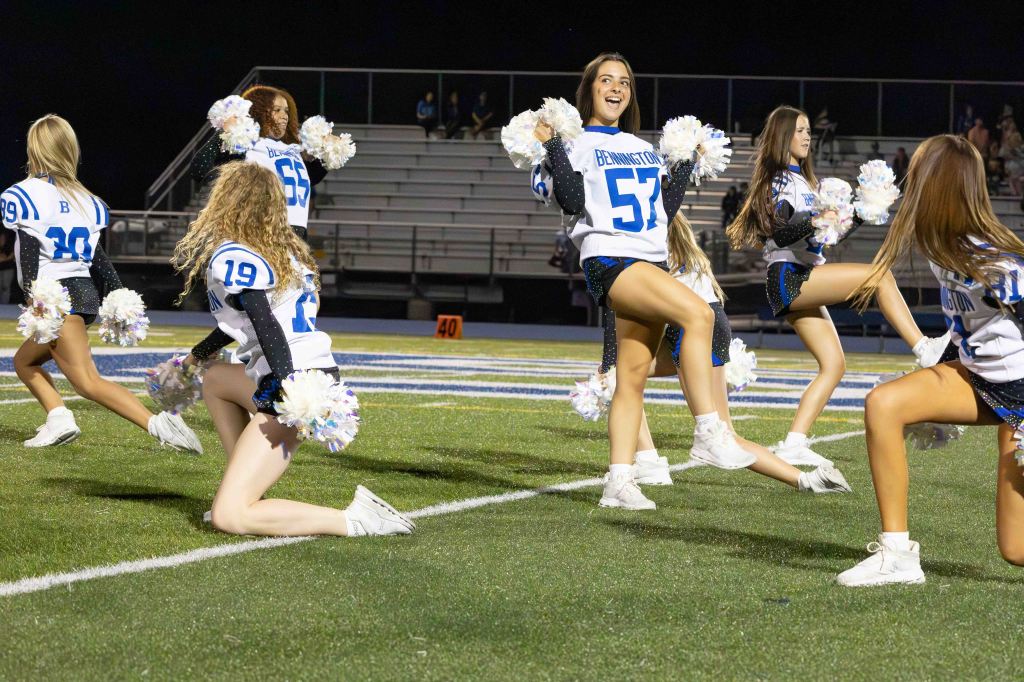 A group of cheerleaders performing a routine on a football field at night, wearing matching uniforms and pom poms.