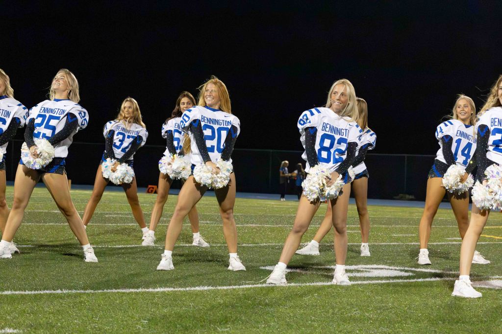 A group of cheerleaders wearing sports jerseys and shorts perform on a football field at night, smiling and holding pom-poms.