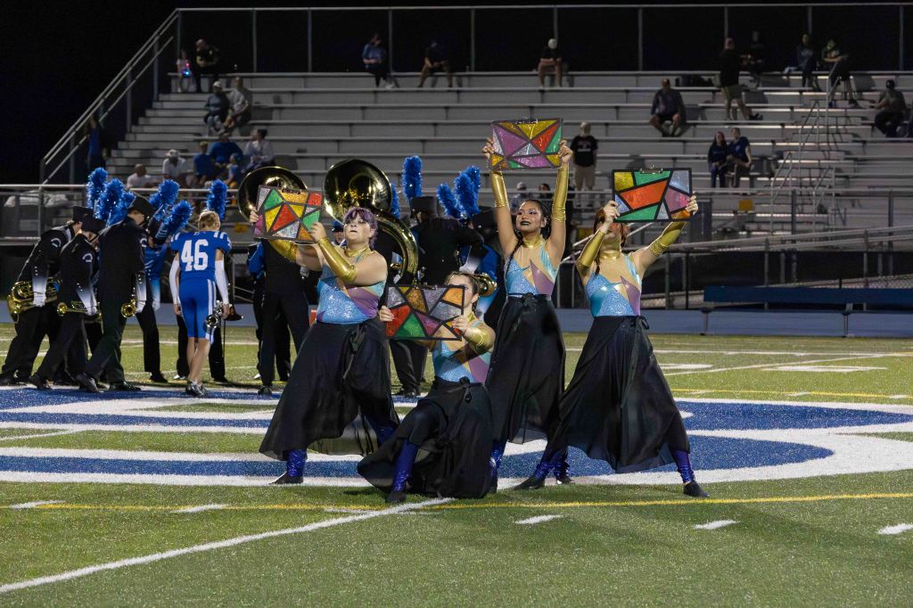 A group of performers in colorful costumes dancing on a football field, holding geometric props while a marching band is visible in the background.