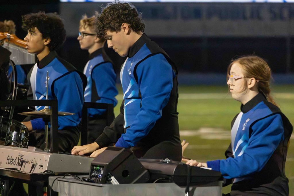 A group of musicians in blue and black uniforms performing on a football field. One musician plays a keyboard while another plays drums, with a focused expression. A young woman in glasses observes the performance.