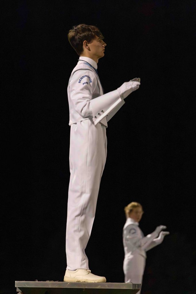 A young male marching band conductor stands on a platform, wearing a white uniform and gloves, with a focused expression. Another band member is visible in the background, also in uniform.