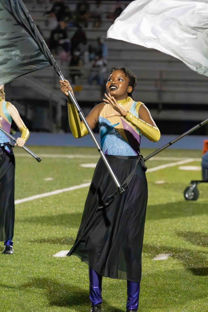 A performer in a colorful costume is joyfully waving a flag during a field show, showing expressive facial gestures.