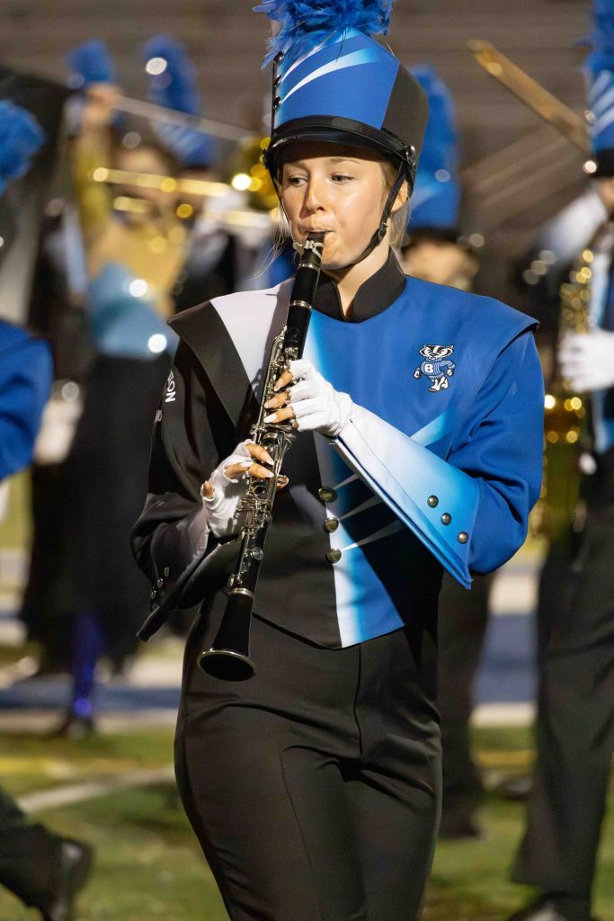 A member of a marching band playing the clarinet, wearing a blue and black uniform with a tall hat and a blue plume, performing on a sports field at night.