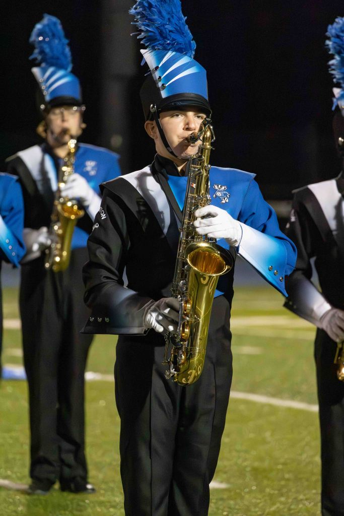 A marching band saxophonist wearing a blue and black uniform with a feathered hat, performing on a football field at night.