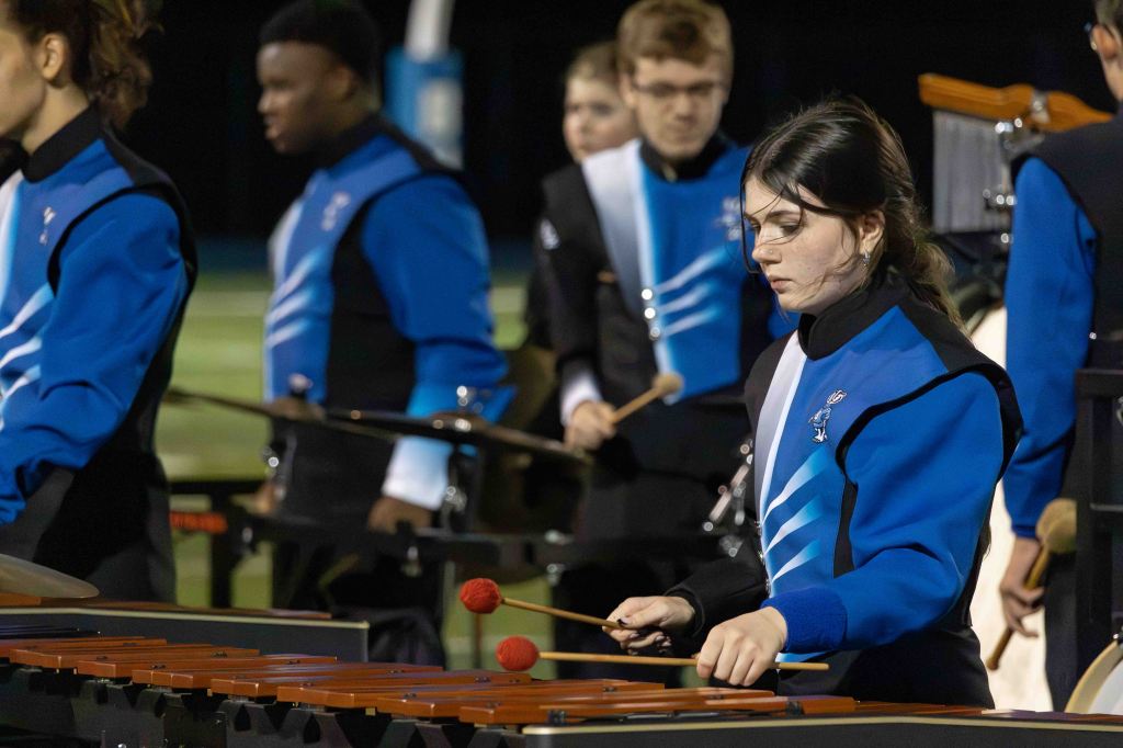 A female percussionist in a blue uniform plays a marimba while other musicians perform in the background on a football field at night.