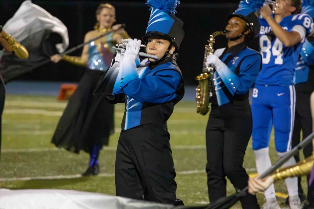 A young musician wearing a marching band uniform plays a flute on a football field, surrounded by fellow band members in various uniforms, including a saxophonist and performers with flags.