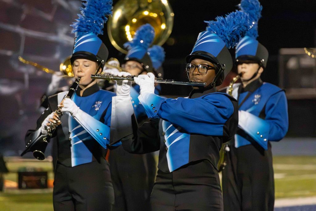 A group of band members in blue and black uniforms performing on a field. One musician is playing the flute while others play different instruments in the background, highlighting their coordinated attire and focus during the performance.