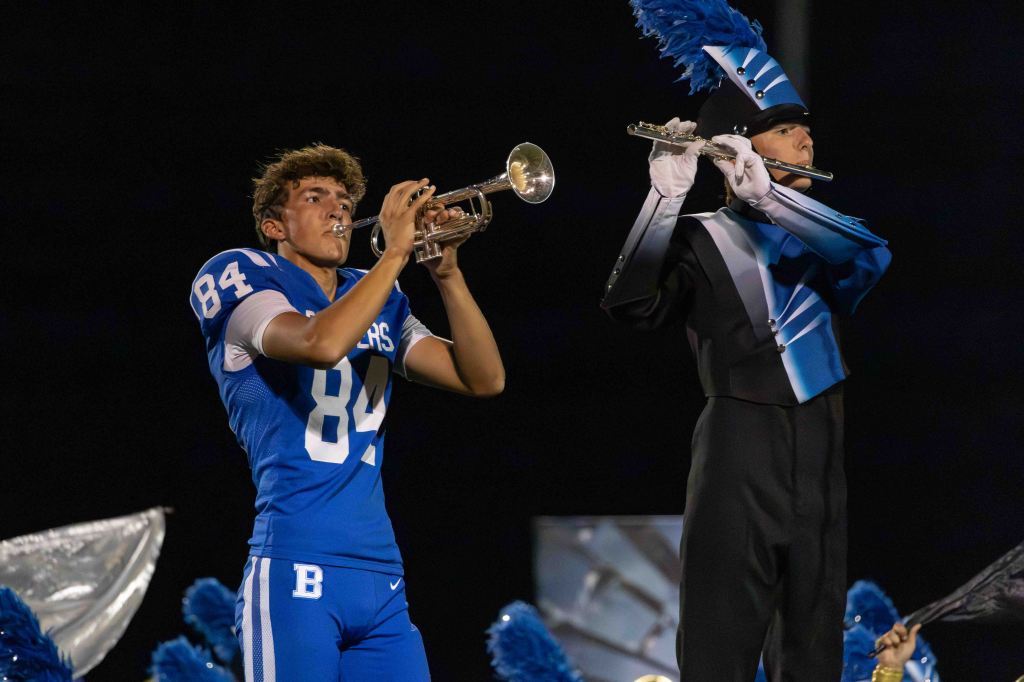 A football player in a blue jersey with the number 84 plays a trumpet while standing next to a marching band member in a black and blue uniform, also playing a trumpet.