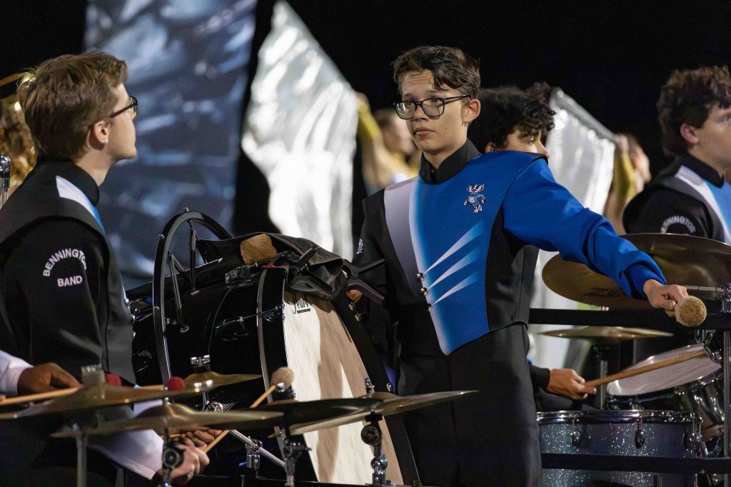 Two young musicians in band uniforms are performing on stage. One musician plays a drum while the other holds a mallet, focused on the performance.