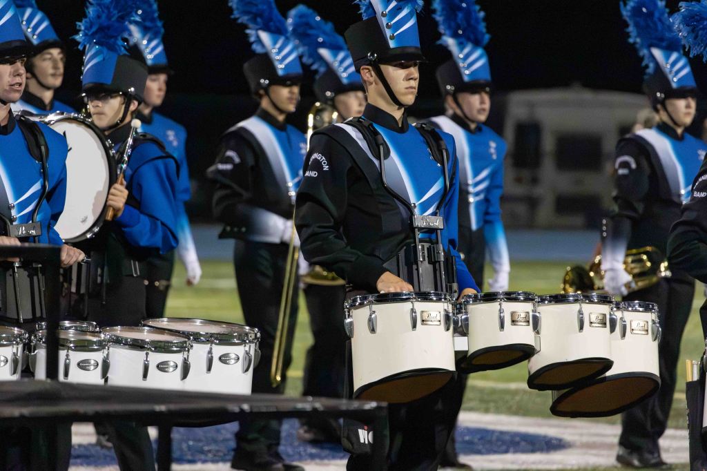 A group of marching band members in blue and black uniforms, standing on a football field at night. The focus is on a drummer wearing a hat and holding a set of white drums.