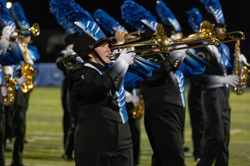 A marching band performing on a field at night, featuring a musician playing the trombone, dressed in a black and blue uniform with feathers on the hat, accompanied by other band members.