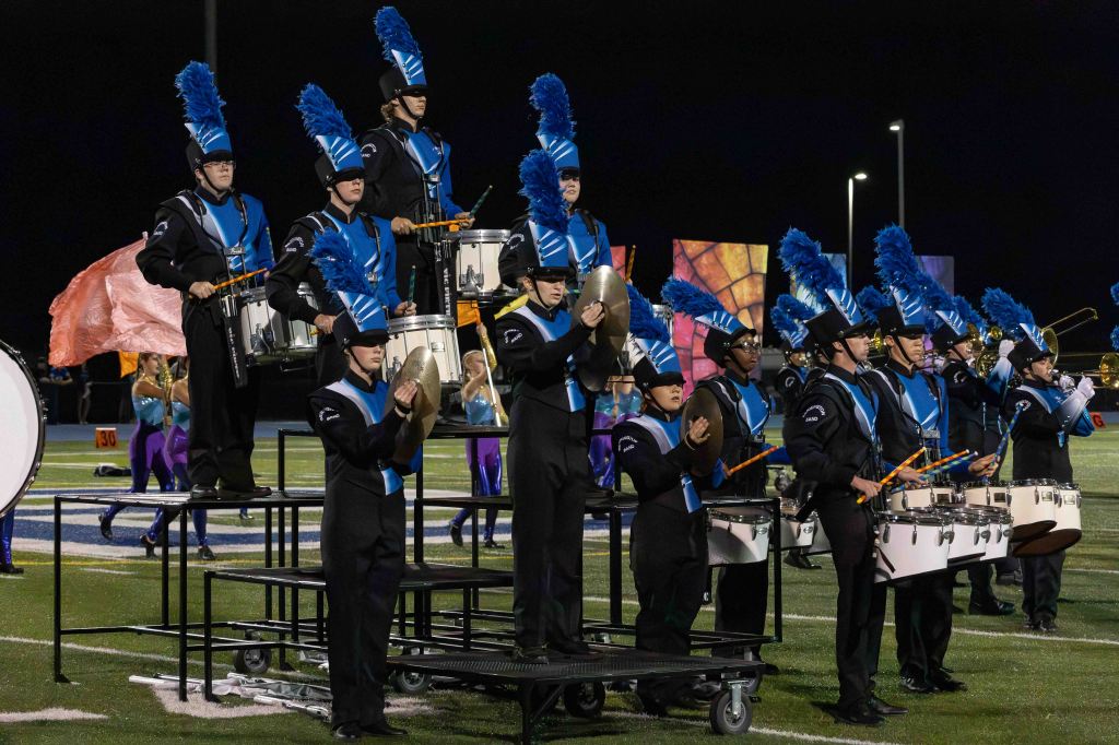 A marching band performs on a field at night, featuring musicians in black uniforms with blue accents and plumes, playing drums and cymbals.