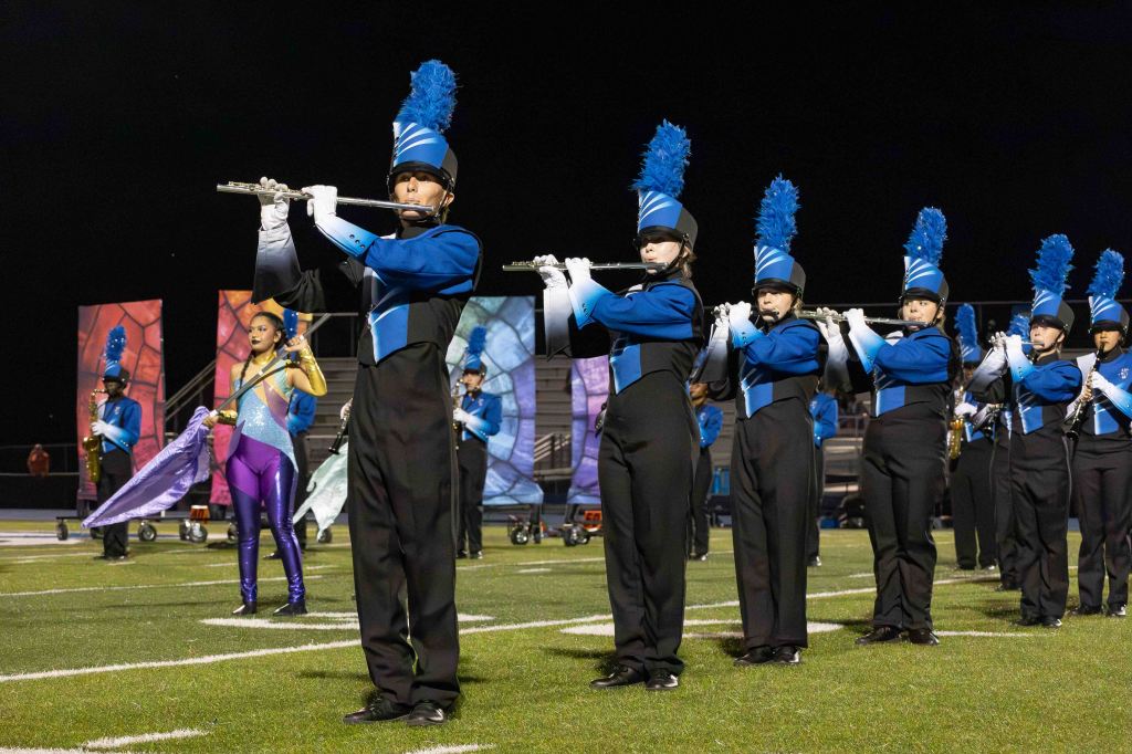 A group of marching band members in blue and black uniforms perform on a football field at night, with a focus on several flutists playing their instruments. Colorful backdrops can be seen in the background.